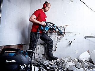 Een man in een rood shirt met gehoorbescherming, die een Makita breekhamer, gekoppeld met een Makita stofzuiger gebruikt